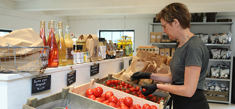 Ett affärsbiträde plockar i färska tomater och grönsaker som står uppställt i en lantbod.